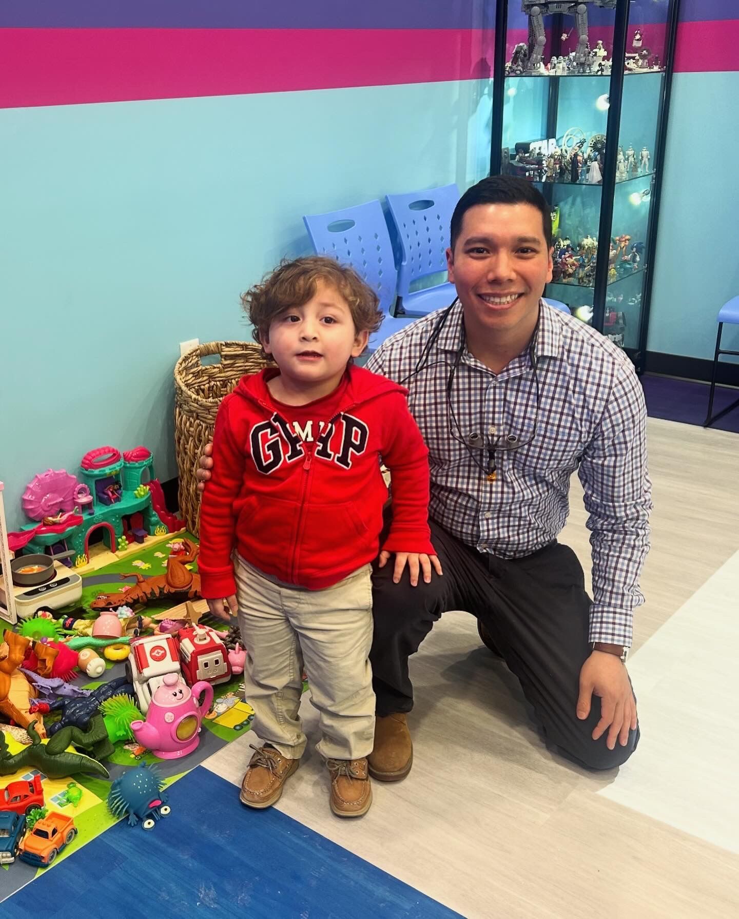 Man and child smiling in a playroom beside a pile of colorful toys.