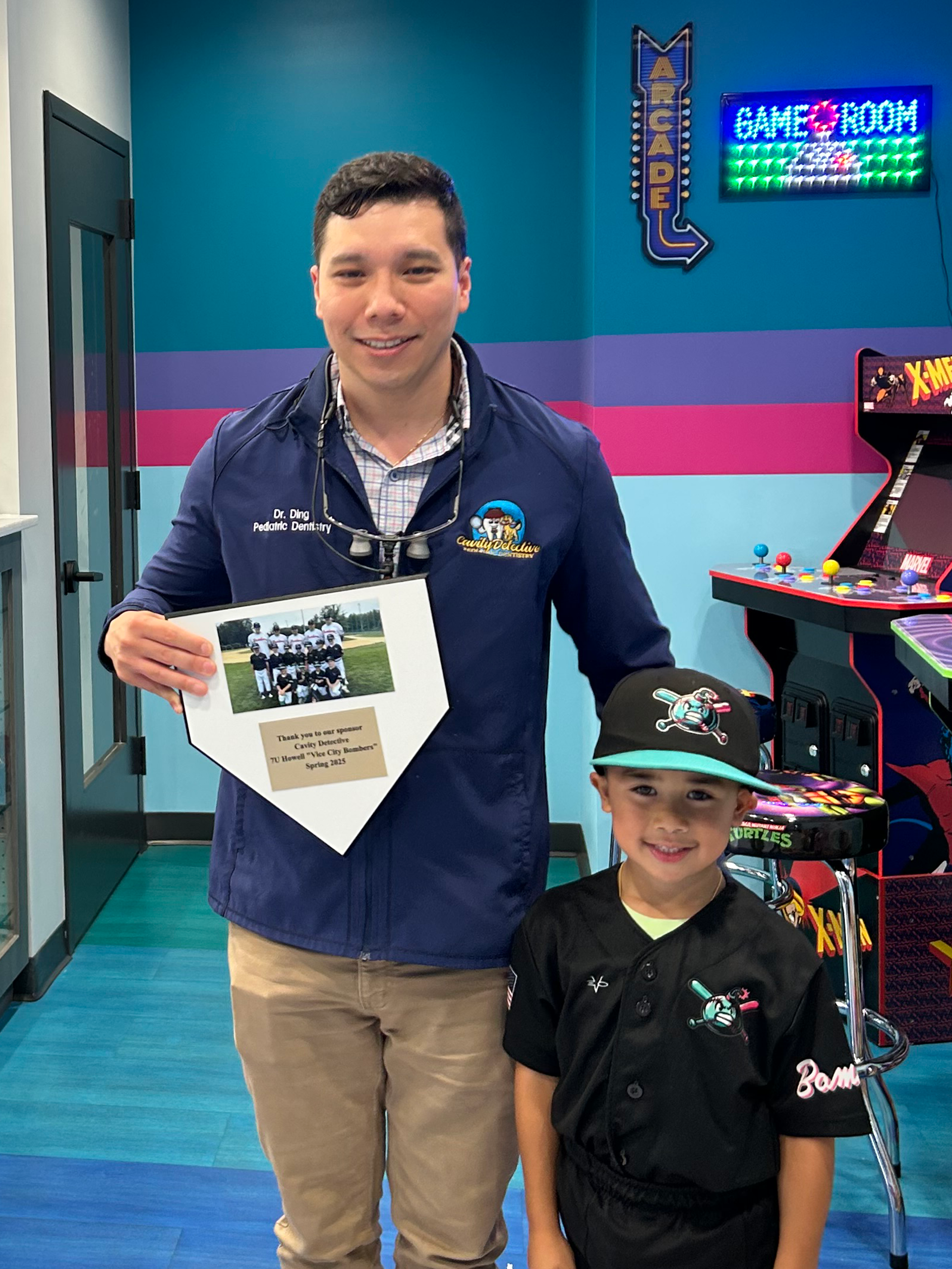 Man in blue shirt and child in black uniform holding a certificate in a colorful arcade room