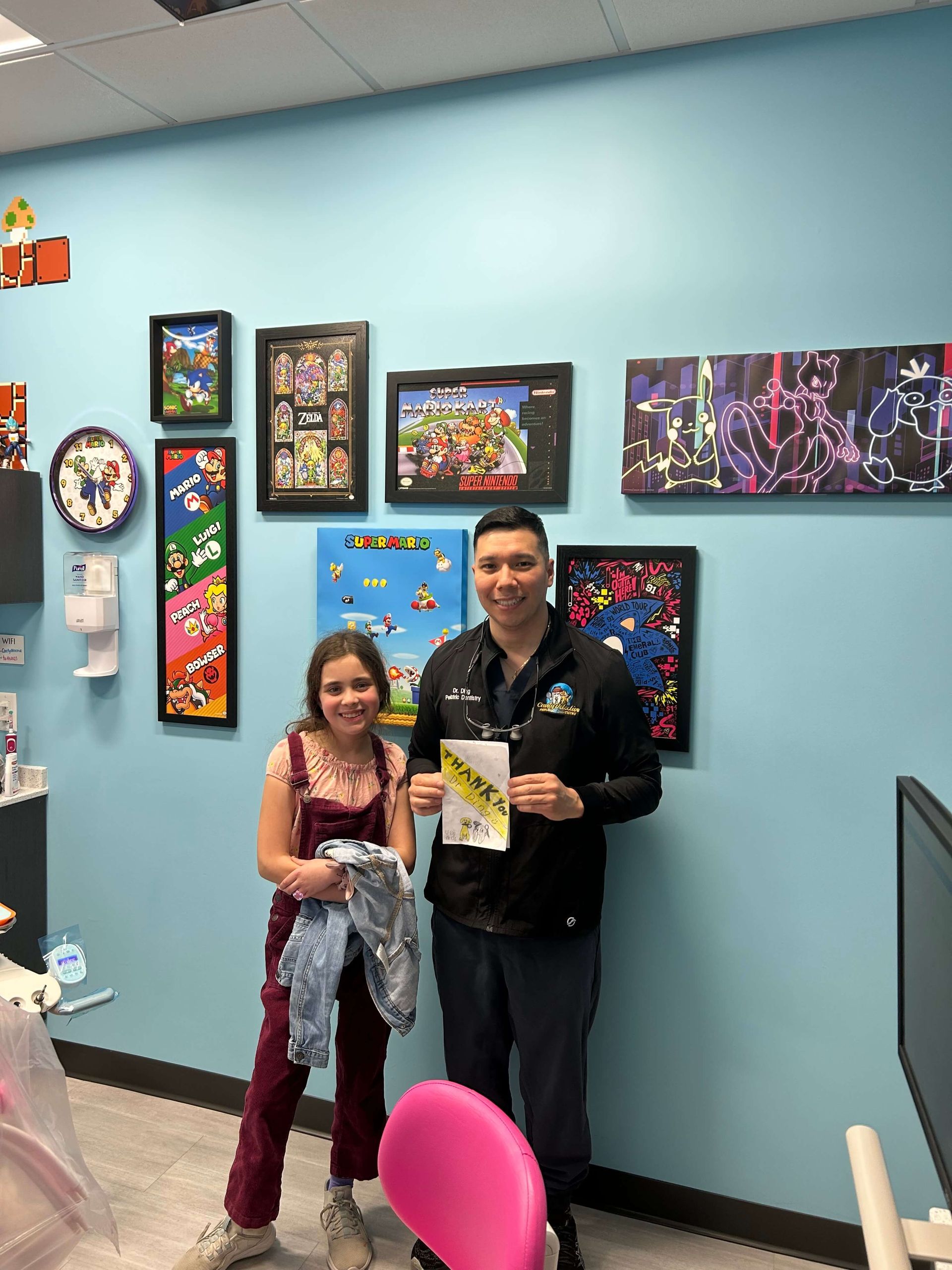 Two people posing in front of a blue wall with colorful framed artwork, holding an item and smiling.