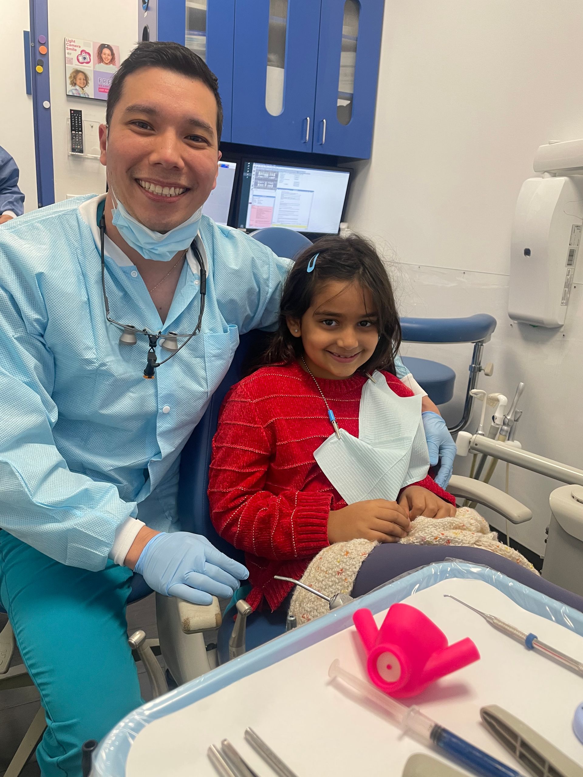 Smiling dental worker with child patient in exam chair, holding a pink toy in a dental office.