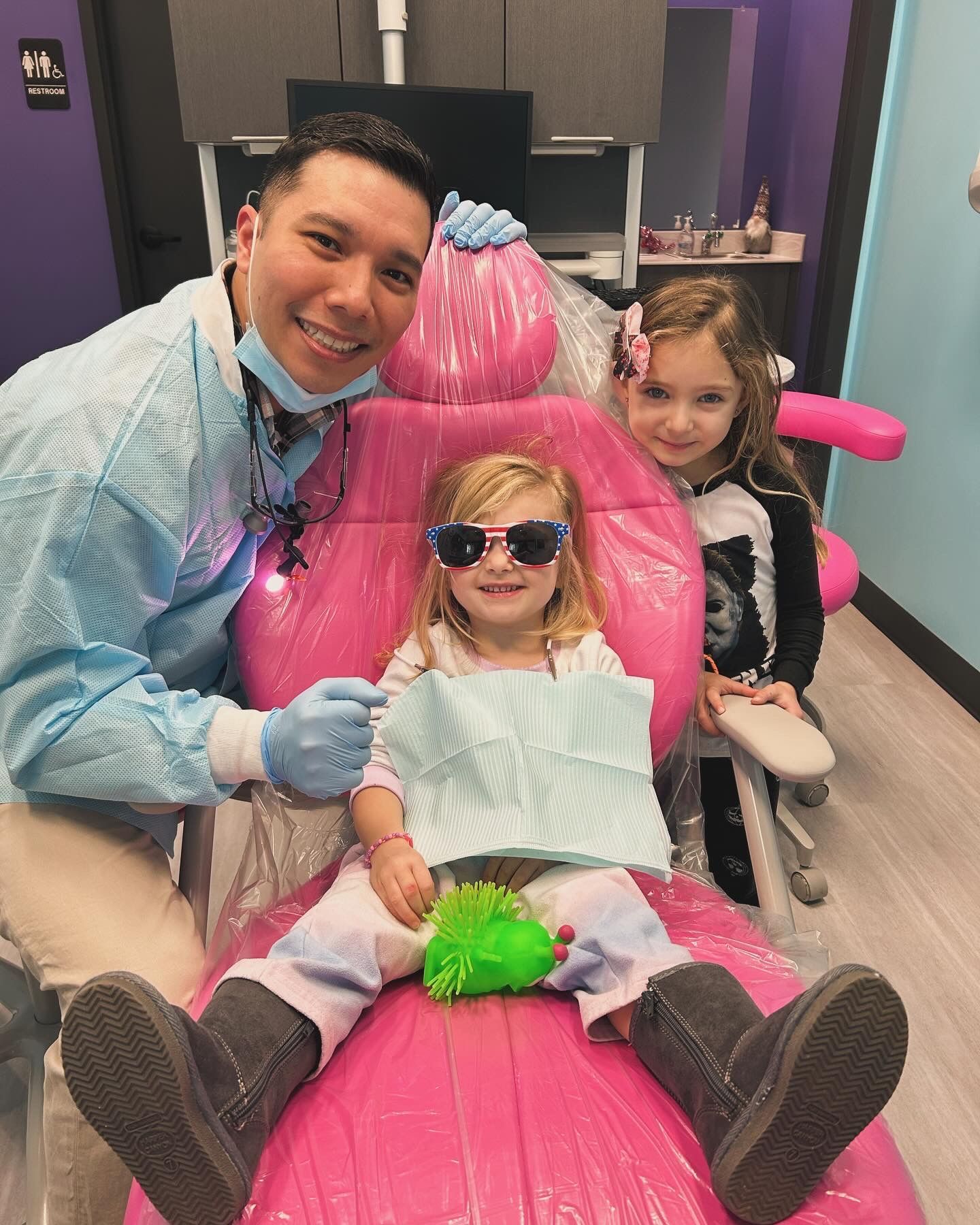 Smiling child in sunglasses sits in a pink salon chair with a man and another child in a salon room